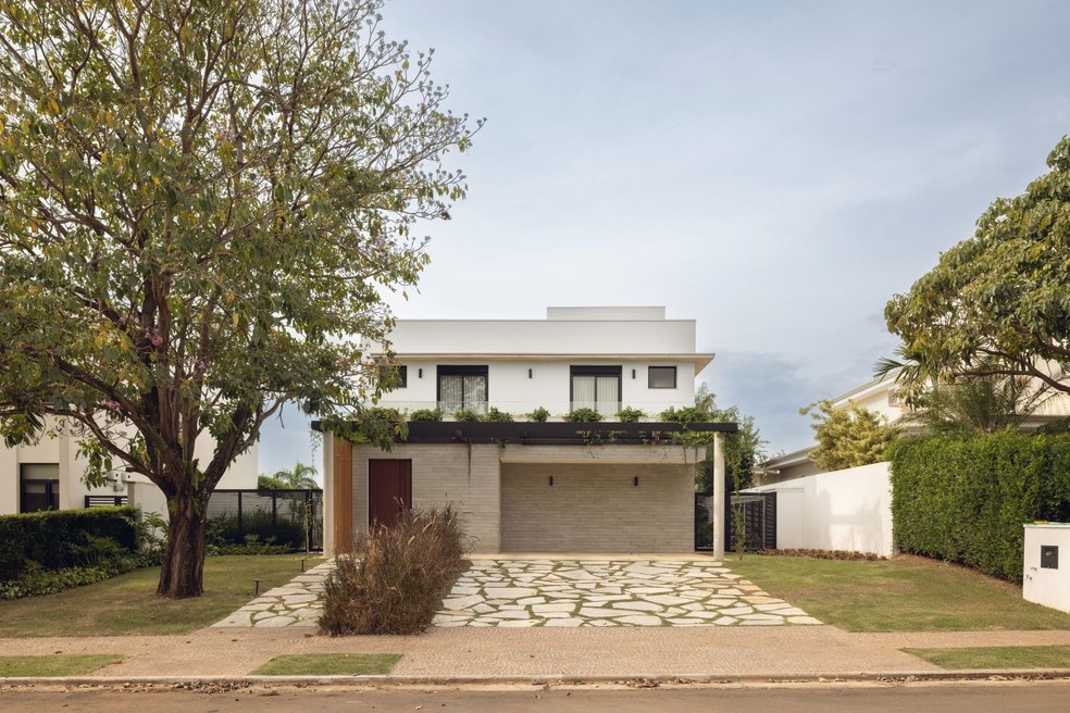 FACHADA | Volumetria limpa e linhas retas marcam a arquitetura da casa, cuja fachada foi revestida de ripas de concreto da Munó no bloco térreo. O paisagismo assinado por Jardineiro Fiel, com execução de Helena Paisagismo, mistura espécies como bromélia e samambaia na varanda, capim-do-texas marcando a entrada, além de grama esmeralda. A grande árvore existente foi mantida — Foto: Maura Mello/Divulgação