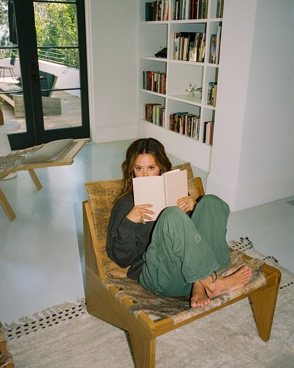 Ashley Tisdale lendo na sua poltrona. Ao fundo é possível ver a sua estante com diversos livros — Foto: Instagram / Reprodução