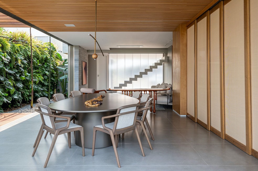 SALA DE JANTAR | A área de refeições, bem como toda a ala social, pode ser separada da cozinha por um painel de correr feito com lâmina natural de freijó e palhinha, executado pela Quéven. O acessório traz privacidade, mas ainda permite certa permeabilidade visual pela presença da palhinha. Mesa de jantar "Abacate", de Roberta Banqueri. Cadeiras "Morris", de Fernando Zanardi. Pendente "Flutua", de André Ferri. Obra de arte sobre a mesa do acervo pessoal dos moradores. Aparador "Chuva", de Leo Romano. Quadro da artista Annike Limborço. Jardim vertical com plantas das espécies samambaia americana, Philodendron cordatum, Philodendron Burle Marxii e Monstera deliciosa mini, executado pela empresa Muro Verde — Foto: Favaro Jr. / Divulgação