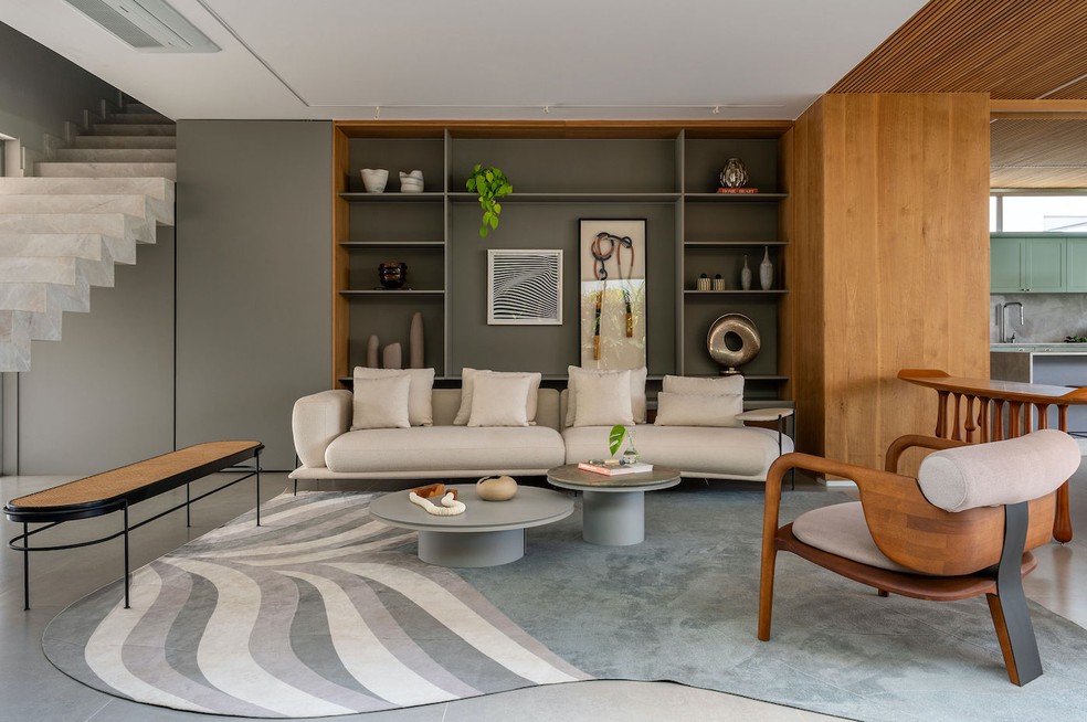 LIVING | A decoração foi pensada com uma paleta neutra com pitadas de tons verdes, como no tapete "Tropical", de Bruno Faucz para a Tapetah. Marcenaria de lâmina natural de freijó e laca cinza, executada pela Quéven. Escada de mármore branco Paraná, executada pela Lopes Mármores. Sofá do acervo pessoal dos moradores. Poltrona "Longeva", de Maurício Bomfim. Mesas de centro "Alma", de Roberta Banqueri. Aparador "Chuva", de Leo Romano. Banco "Iaiá", de Gustavo Bittencourt. Tapete Tropical, de Bruno Faucz para a Tapetah. Os objetos decorativos da estante são das lojas Ideias On e Degusta. Vaso com jibóia pendente. Iluminação técnica da Arteluz Piracicaba — Foto: Favaro Jr. / Divulgação