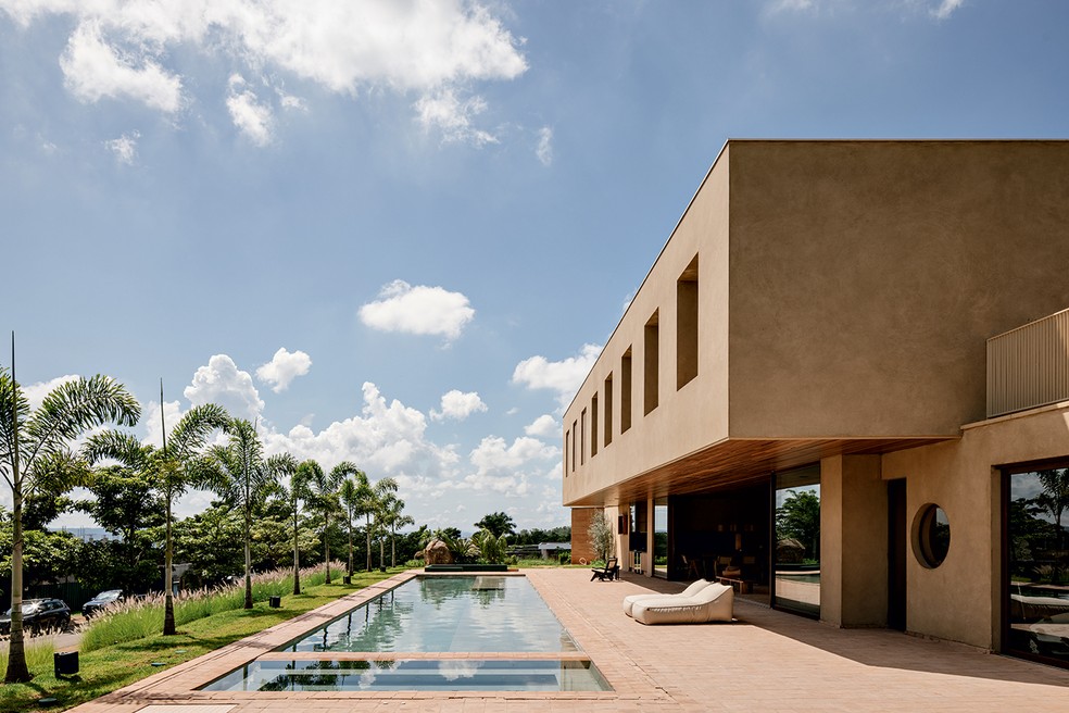 ÁREA EXTERNA | O volume do pavimento superior avança 3 m em balanço e forma a grande varanda voltada para a piscina, revestida de placas cimentícias cor cinza da Tresuno. As mesmas placas em tom terroso cobrem todos os pisos das áreas externa e interna, integradas com a abertura de todo o vão pelos painéis deslizantes de vidro da Conceito Esquadrias de Alumínio. Poltronas da Casa Atica — Foto: Fran Parente / Divulgação