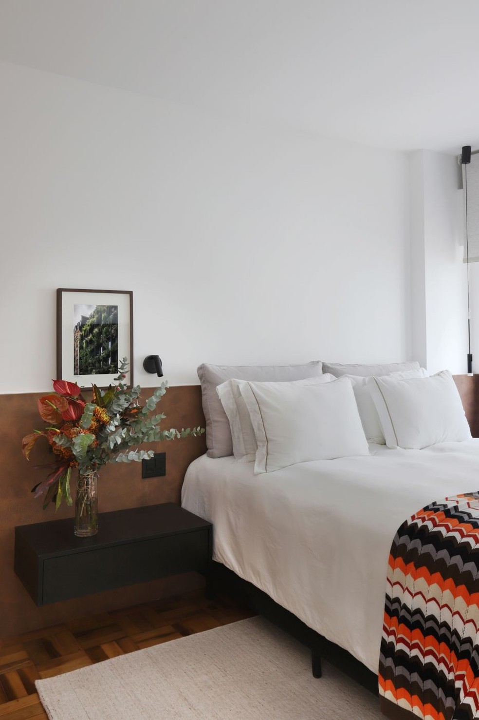 SUÍTE | O painel da cabeceira, com acabamento que lembra aço corten, e a mesa lateral suspensa foram executados pela AMA Marcenaria. Roupa de cama da Testani Casa. Sobre o piso de taco de perobinha, tapete da by Kamy. Arranjo feito pelo arquiteto com antúrios e eucaliptos — Foto: Mariana Orsi/Divulgação