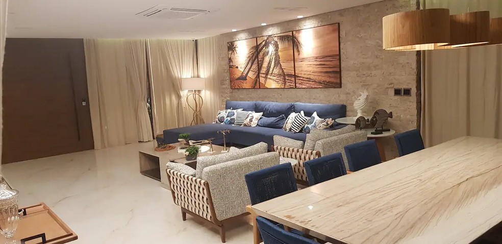 A sala de estar tem um sofá azul bem grande e um quadro na parede com a imagem de uma praia — Foto: Airbnb / Reprodução