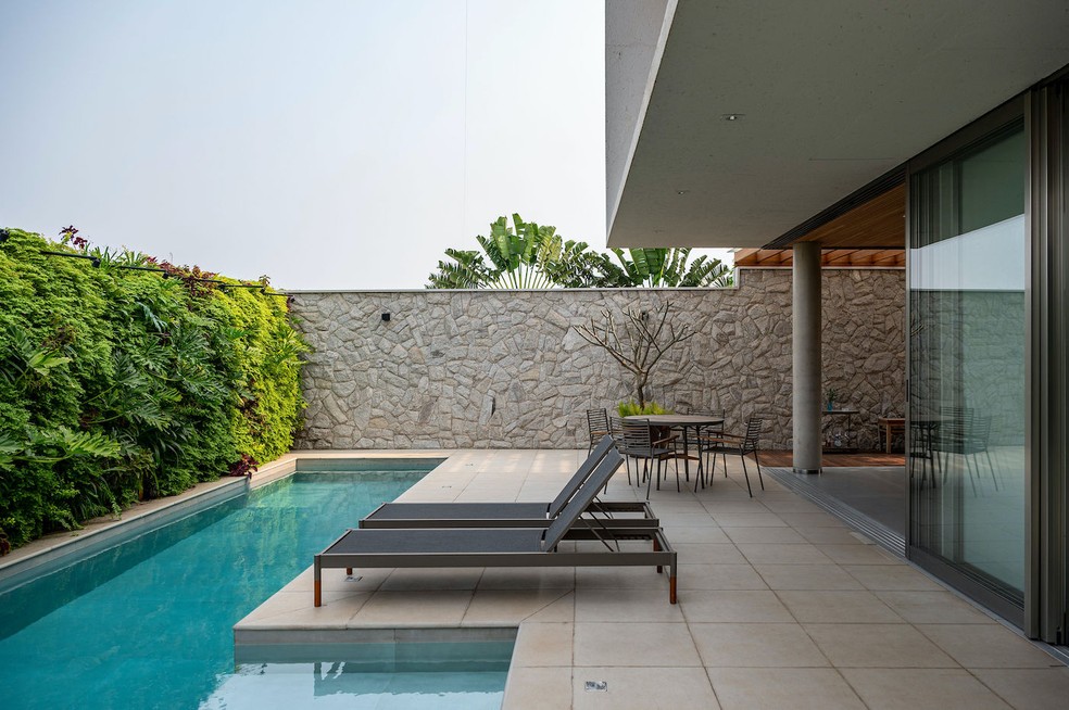 PISCINA | Aos fundos do terreno, o projeto trouxe uma piscina, que ganhou a companhia de um grande jardim vertical com plantas das espécies guaimbê, Philodendron undulatum, brilhantina, cóleus, fitônia e agapanto, executado pela empresa Muro Verde. Piso de revestimento Lime Fendi, da Castelatto. Muro de pedra moledo fornecida pela Mardois Pedras e executada por profissional local. A piscina recebeu os revestimentos Prainha em Lime Fendi, da Catelatto, e porcelanato Make Corda, da Gardenia. Espreguiçadeiras modelo “Skyfolding”, da Dona Flor. Mesa com cadeiras “Lira”, da Mestre Artesão — Foto: Favaro Jr. / Divulgação