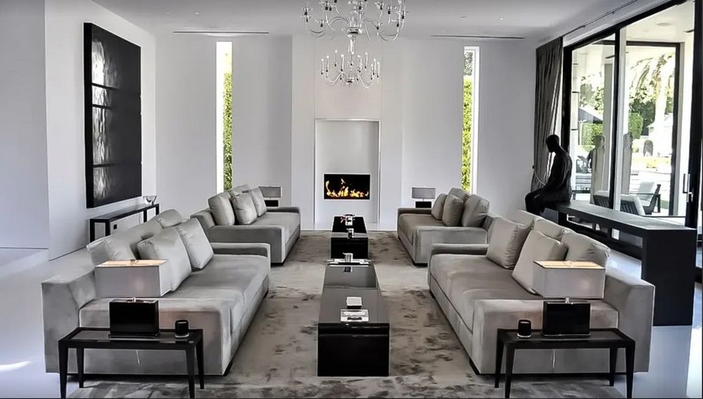 A sala de estar tem lareira, um lustre e diversos sofás em tons claros — Foto: Robb Report / Reprodução