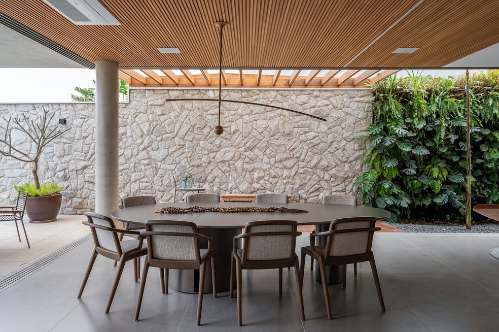 SALA DE JANTAR | Quando as portas de vidro do chão ao teto são abertas, a sala de jantar fica totalmente integrada com a área externa, trazendo amplitude. Mesa de jantar "Abacate", de Roberta Banqueri. Cadeiras "Morris", de Fernando Zanardi. Pendente "Flutua", de André Ferri. Obra de arte sobre a mesa do acervo pessoal dos moradores. Forro ripado de madeira tauari, da Indusparquet. Muro de pedra moledo fornecida pela Mardois Pedras e executada por profissional local. Pergolado feito de madeira freijó por empresa local. Vaso com árvore jasmim-manga, com forração de aspargo real. Jardim vertical com plantas das espécies samambaia americana, Philodendron cordatum, Philodendron Burle Marxii e Monstera deliciosa mini, executado pela empresa Muro Verde — Foto: Favaro Jr. / Divulgação