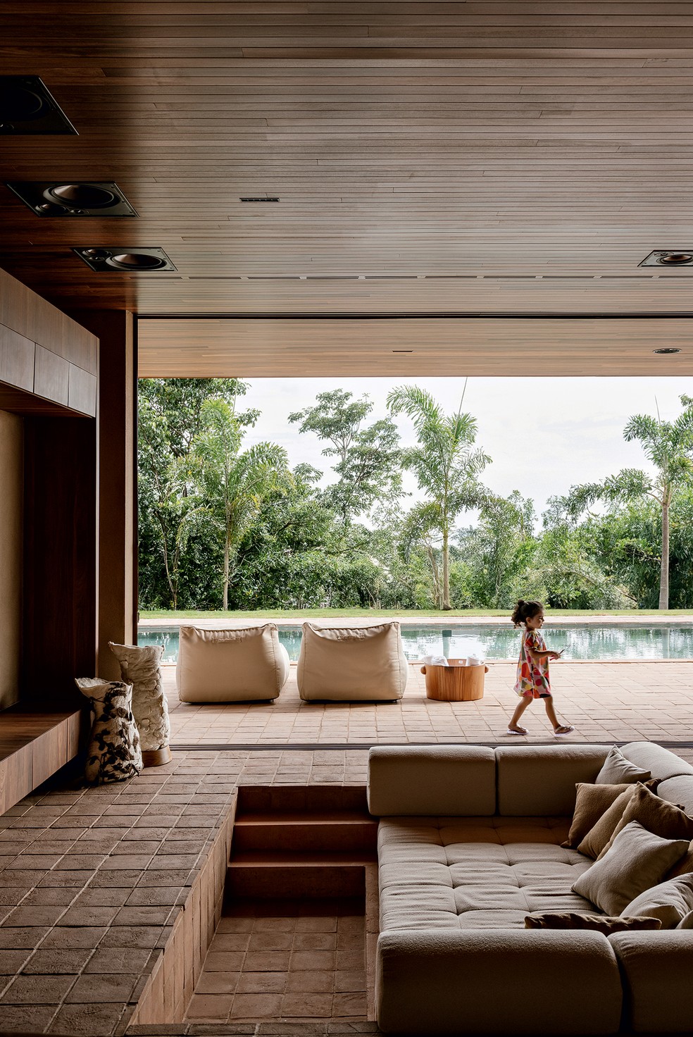 ÁREA SOCIAL | A filha do casal está na área externa totalmente integrada ao living. Para a TV ser vista de todos os cantos, a sala tem sofá negativo projetado pelos arquitetos no nível mais baixo do piso de placas cimentícias da Tresuno — Foto: Fran Parente / Divulgação