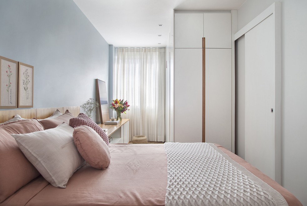 O quarto ganhou uma atmosfera tranquila com a parede azul e a roupa de cama rosa. Projeto do escritório Ateliê Concreto — Foto: Raiana Medina / Divulgação