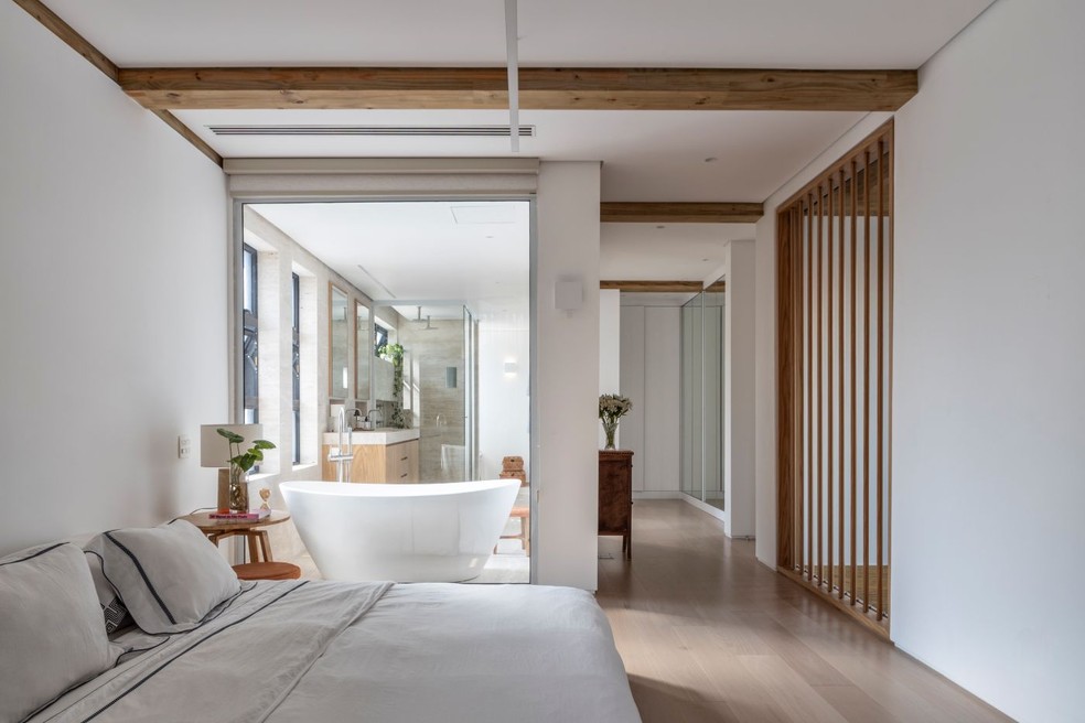QUARTO CASAL | A paleta clara domina o ambiente, aquecido pela madeira presente na estrutura e no piso da Parket. Mesa de canto e objetos da dpot. Ao fundo, a sala de banho com banheira fica à mostra através da parede de vidro — Foto: Ruy Teixeira/Divulgação