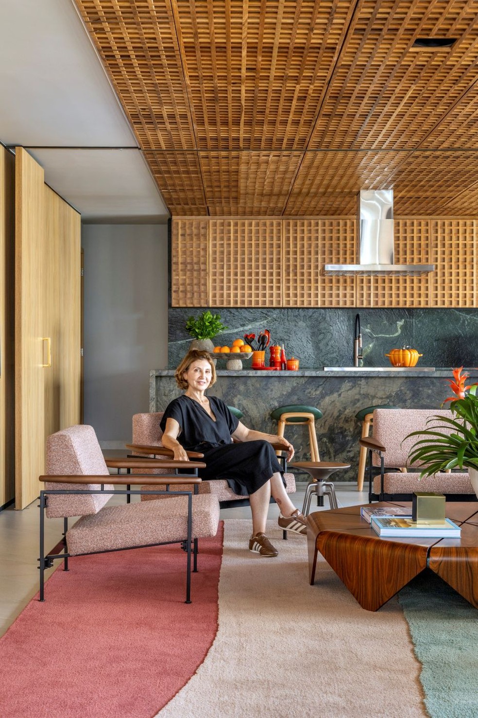 RETRATO | A moradora Rosângela Santana está na área social, tendo ao fundo a cozinha, com painel de muxarabi e marcenaria desenhados pelo Plano Studio e executados pela movelaria Casa Marista. O espaço combina ainda quartzito Vitória Régia nas bancadas principais e quartzito Bronzita na ilha, ambas executados pela Gruta Mármores. Banquetas Joy, de Jader Almeida, na Via Condotti — Foto: Edgar Cesar/Divulgação