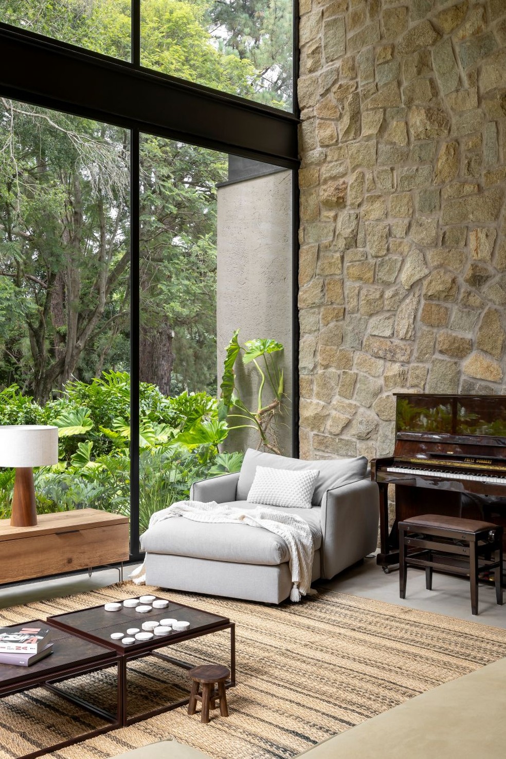 SALA DE ESTAR | Materiais naturais, como a parede de pedra modelo e o piso de cimento queimado, criam uma estética rústica. Chaise cinza, da Artefacto, ao lado do piano — Foto: Keniche Santos/Divulgação