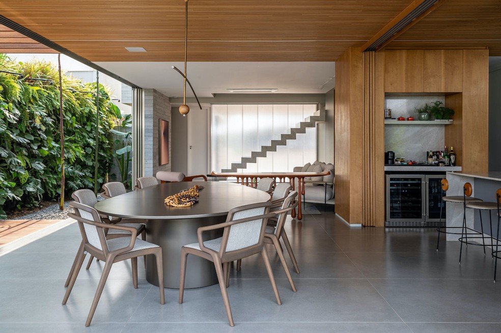 SALA DE JANTAR | Integrado à cozinha e à sala de estar, o espaço de refeições faz conexão também com a área externa. Ao fundo, o painel translúcido de policarbonato estrutural, da Arkos, traz luminosidade à ala social. Mesa de jantar "Abacate", de Roberta Banqueri. Cadeiras "Morris", de Fernando Zanardi. Pendente "Flutua", de André Ferri. Obra de arte sobre a mesa do acervo pessoal dos moradores. Marcenaria de lâmina natural de freijó e laca cinza, feita pela Quéven. Ao lado, o nicho revestido de mármore branco Paraná, executado pela Lopes Mármores, deu lugar a um cantinho de café e acomoda as adegas da Evol. Piso de porcelanato Full Concret, da Roca. Forro ripado de madeira tauari, da Indusparquet — Foto: Favaro Jr. / Divulgação