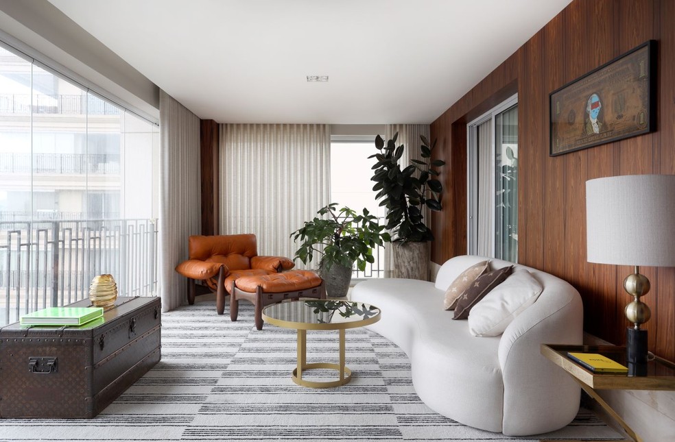 O sofá Curvo, do Estúdio Lider, traz linhas curvas para o ambiente, que ganhou o aconchego do tom alaranjado da poltrona Mole, de Sergio Rodrigues, na Dpot. Mesa de centro de ferro e vidro, da Cápsula Decor, que também forneceu a mesa lateral Z e o abajur. No painel de lâmina de pau-ferro, está o quadro assinado por Cranio, na B.I.A Colecionáveis. Sobre o tapete da by Kamy, baú da Louis Vuitton garimpado pelos moradores. Cortinas de gaze de linho fornecidas pela Casa Fortaleza. Vasos e plantas são da Blooms Floral Design by Tomaz Pinto — Foto: Marco Antonio/Divulgação | Projeto do arquiteto Robert Robl