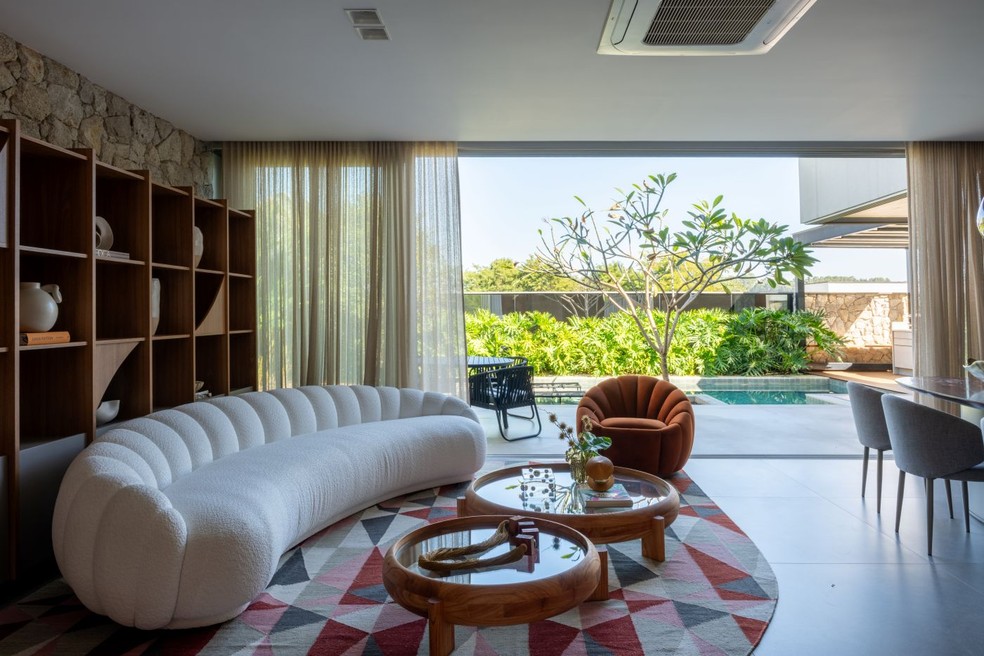 LIVING INTEGRADO | A reforma da casa de 334 m² valorizou a convivência por meio de ambientes sociais integrados em conexão com a área externa. Ao fundo, o muro de vidro espelhado amplia a percepção espacial e reflete o paisagismo idealizado e executado por Spagnhol Plantas Ornamentais, com o maciço de guaimbê ao longo da borda da piscina — Foto: Junior Favaro/Divulgação