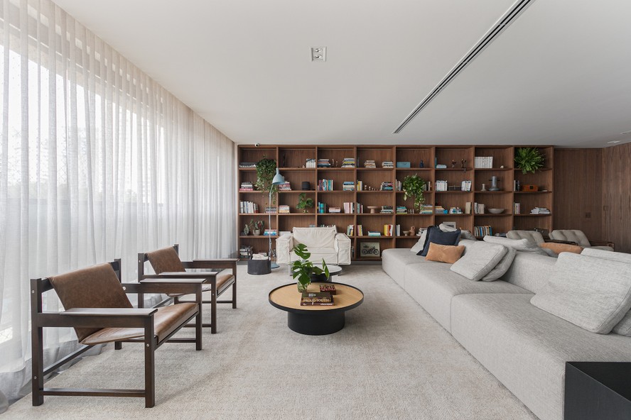 LIVING | A grande estante do piso ao teto, executada pela Affatto Móveis em folha natural de nogueira, organiza livros dos moradores e objetos da história deles. Sofá em ilha 'MCS-65', da Micasa. Poltronas 'Lia', de Sergio Rodrigues. Ao fundo, poltrona 'Less', do estudiobola. Luminária de piso azul da Lumini. Mesas de centros feitas pela Affatto Móveis e Rocamar Marmoraria em serralheria com acabamento em preto microtexturizado. Tapete da By Kamy. Cesto preto e almofadas do acervo dos moradores. Cortina em linho da Tec&Paper. Plantas das espécies samambaia, jibóia, dinheiro em penca e costela-de-adão