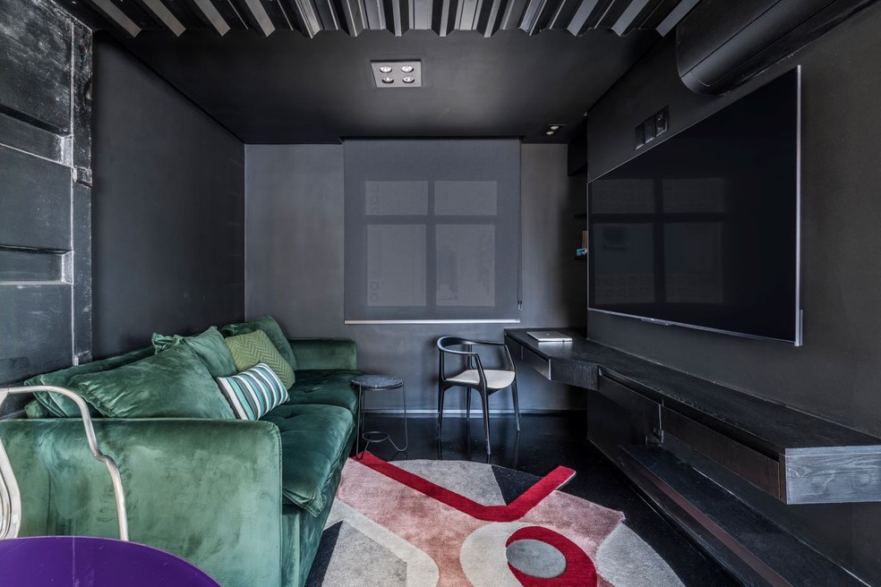 SALA DE TV | A base preta envolve forro de telha metálica e pintura eletrostática, parede lateral recoberta com porta de contêiner reaproveitado, além da marcenaria executada com MDF Preto Absoluto, da Duratex, pela MPA Rocha Marcenaria. As cores ficam por conta do sofá de veludo verde da Constantine Móveis, e do tapete Nova Augusta, desenhado por Ricardo Abreu Arquitetos para By Kamy Verde. Cadeira da Dunelli — Foto: Renato Navarro / Divulgação