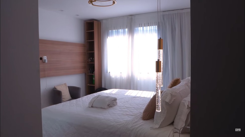 O quarto tem uma decoração minimalista e um painel de madeira em uma das paredes — Foto: Youtube / Reprodução