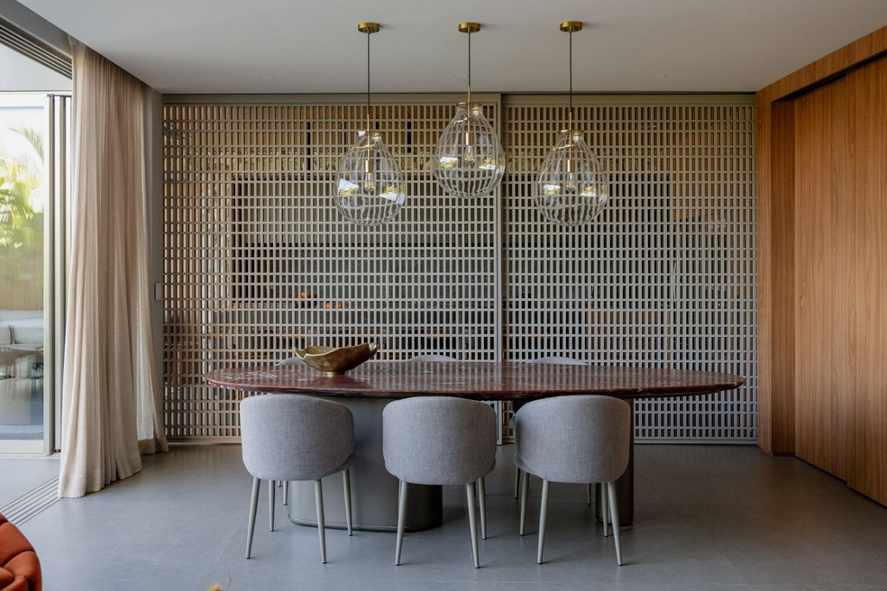 SALA DE JANTAR | A mesa de jantar, da Lider Interiores, com tampo de quartzito Napoleon Bordeaux, da Michelangelo Mármores, foi composta com cadeiras da Escal, na loja Artzzi, estofadas em linho, além de luminárias pendentes de vidro, que valorizam o conjunto e o pé-direito. À frente, os painéis deslizantes de muxarabi com acabamento de laca, desenho exclusivo do escritório, delimita a área gourmet e promovem flexibilidade de uso e controle visual entre os espaços. À direita, os painéis deslizantes em madeira possibilitam o fechamento da sala de TV. Piso de porcelanato da marca Roca, adquirido na loja Spazio Pisos e Revestimentos — Foto: Junior Favaro/Divulgação