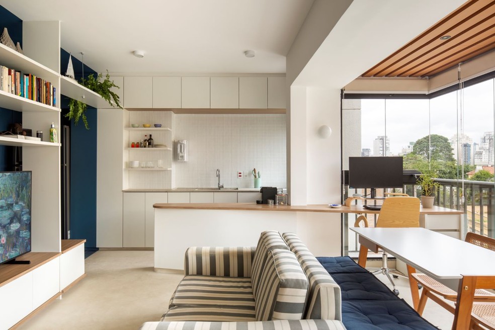 O sofá listrado, em tons neutros, estabelece diálogo com o azul dos futons e com a pintura da parede. Ao mesmo tempo, conversa com a cozinha branca — Foto: Carolina Lacaz/Divulgação | Projeto dos arquitetos Raphael Souza e Daniel Zahoul