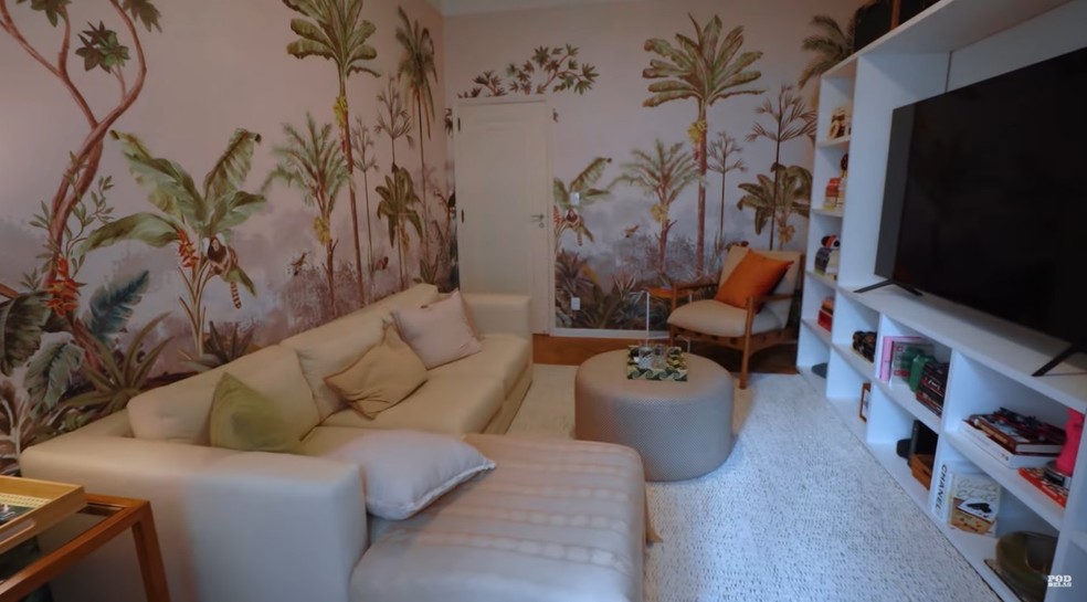 A sala de TV tem um papel de parede de uma floreta, um sofá branco e um móvel com itens de decoração — Foto: Youtube / Reprodução