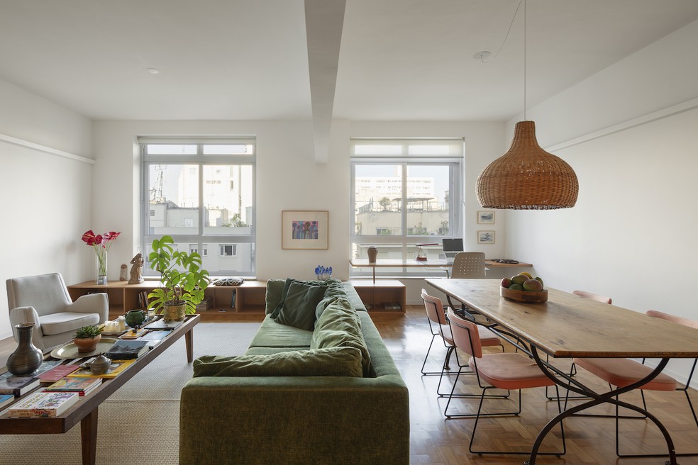 A sala integrada traz mesa de centro feita com estrutura de cama de solteiro de São João Del Rei (MG), que pertencia à avó do morador. Mesa de jantar, cadeiras, tapete e objetos decorativos do acervo do morador. Poltrona "Pé Palito", de Carlo Hauner. Iluminação com fitas da MisterLED inseridas em serralheria produzida por Raul Furlan. Ao fundo, antúrios vermelhos e, na mesa, costela-de-adão. Marcenaria de folha de curupixá, executada pela Marcenaria Quadrato. Projeto dos arquitetos Rosário Pinho e André Scarpa — Foto: André Scarpa/Divulgação