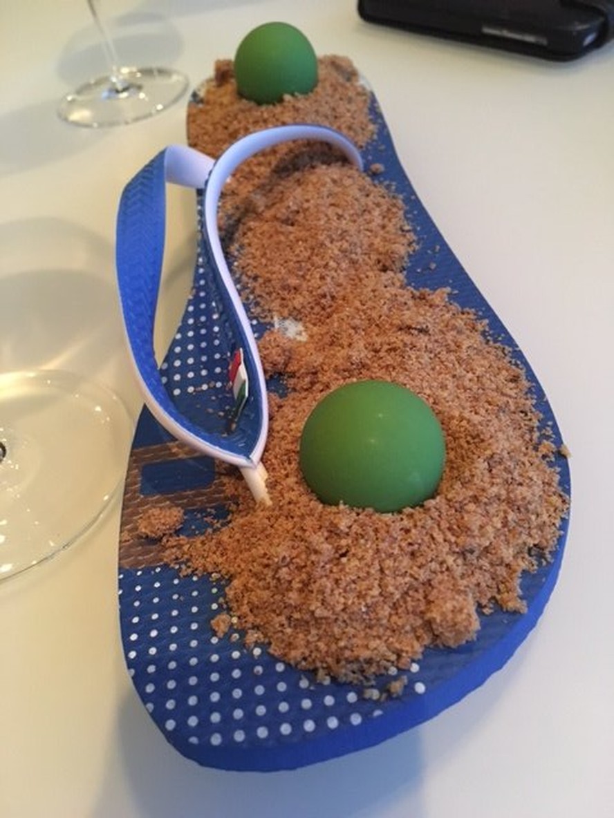 A esfera de chocolate é servida sobre uma cama de farofa de nozes, e o chinelo serve como 'prato'