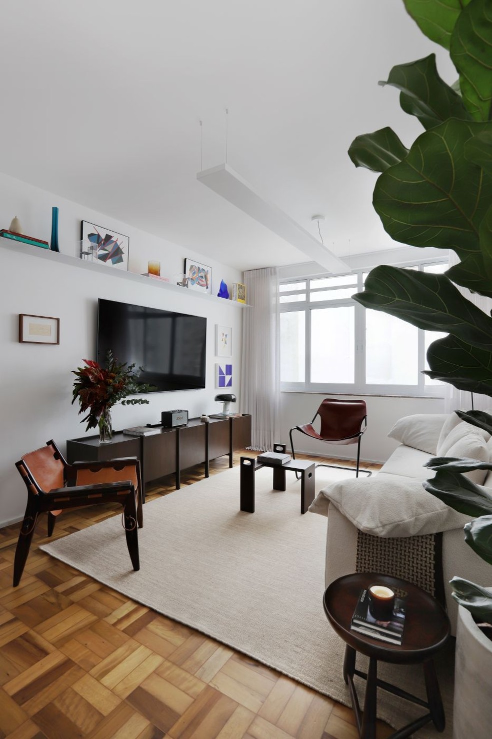 SALA DE ESTAR | O ambiente é composto por aparador Palafita, assinado por Felipe Zorzeto, fazendo as vezes de rack sob a TV, rodeado de obras de arte de nomes como Alfredo Volpi, Almicar de Castro e Marília Bulhões. O design brasileiro é representado ainda por peças como a poltrona Kilin, de Sergio Rodrigues, que também assina o banco Mocho, ao lado do sofá da Più Mobile. Ao fundo, poltrona Paulistano, de Paulo Mendes da Rocha, e, no centro, a peça Banquinho, de Claudia Moreira Salles. Cortinas da Hunter Douglas. Tapete da by Kamy. Luminárias da Dessine — Foto: Mariana Orsi/Divulgação